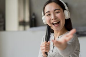 A smiling person wearing headphones sings into a hairbrush, extending their hand forward in a casual indoor setting—capturing the fun of vocal lessons Singapore has to offer.