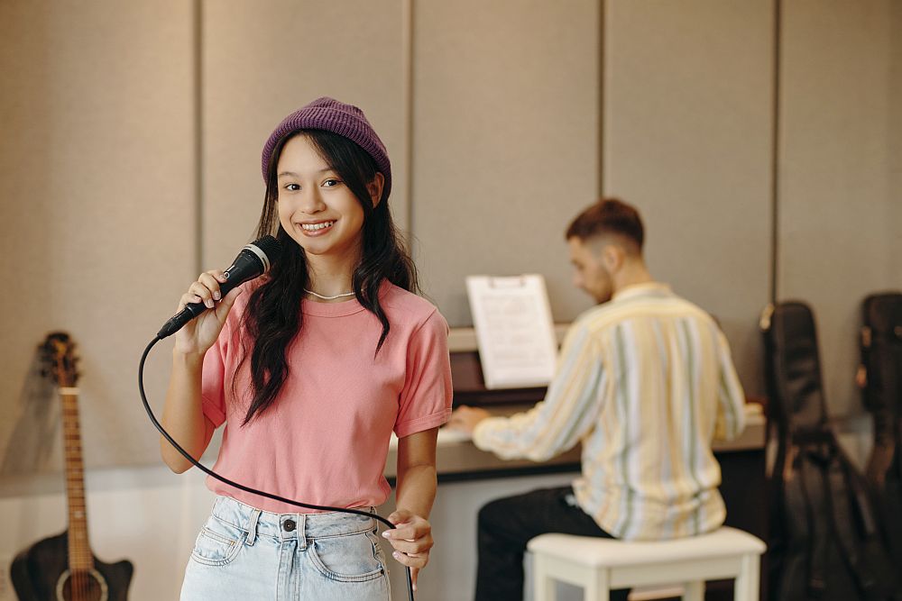 A young woman holds a microphone and smiles at the camera while a man plays the piano in the background of a music studio, capturing the vibrant atmosphere of vocal lessons Singapore.