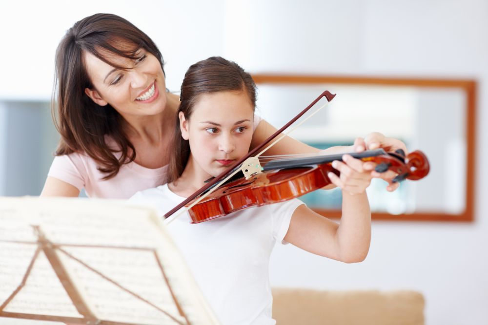 An adult woman helps a young girl play the violin, guiding her posture as they look at sheet music in a bright room, capturing the supportive spirit of violin lessons Singapore is known for.