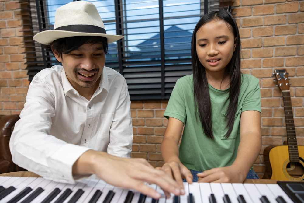 An adult wearing a hat and a child sit at a keyboard piano. As the adult points to the keys, the child looks on—offering a glimpse into what to expect before enrolling in lessons or researching piano lesson fees Singapore. A guitar leans against the wall nearby.