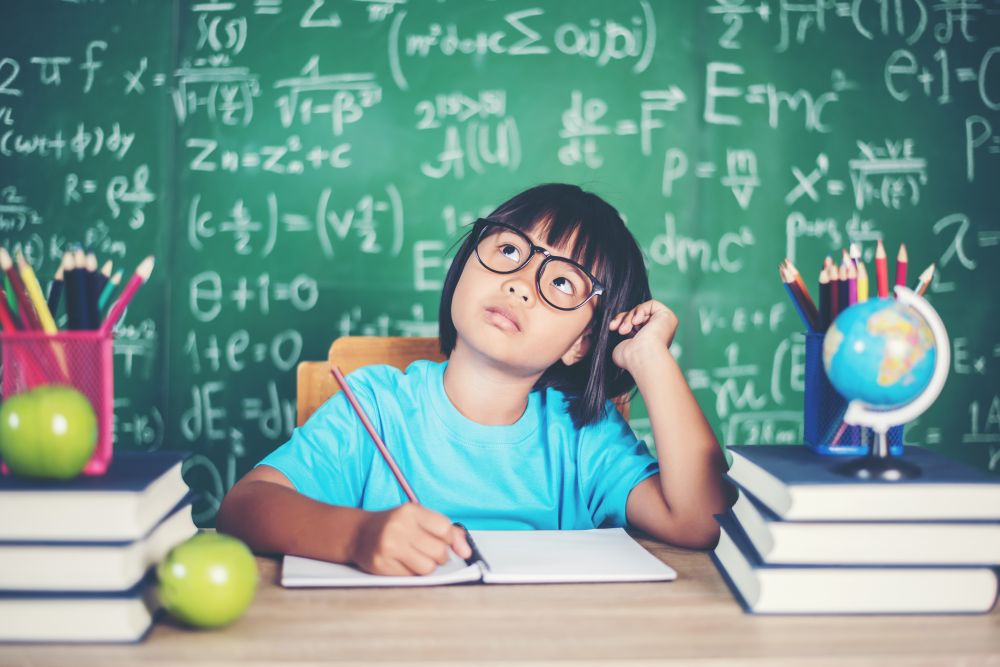 A child in glasses sits at a desk with books and school supplies, writing in a notebook, with a chalkboard covered in math equations in the background—perfectly capturing the essence of Sec 1 Math: What Students Should Focus On First as they begin their learning journey.