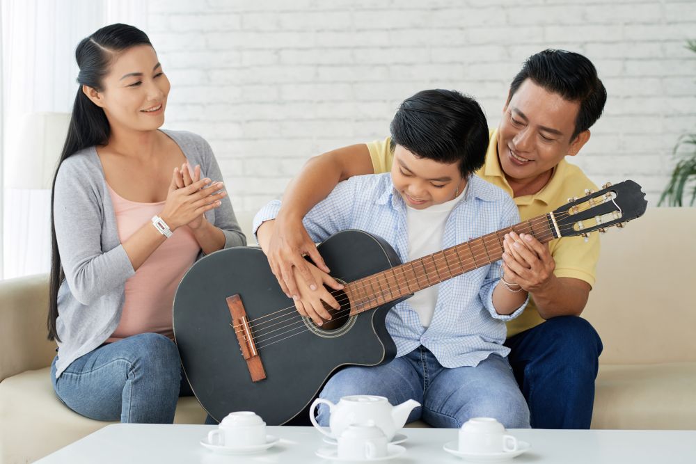 A father and mother sit on a sofa watching their son play an acoustic guitar, a scene reflective of what parents should know about singing classes for kids.